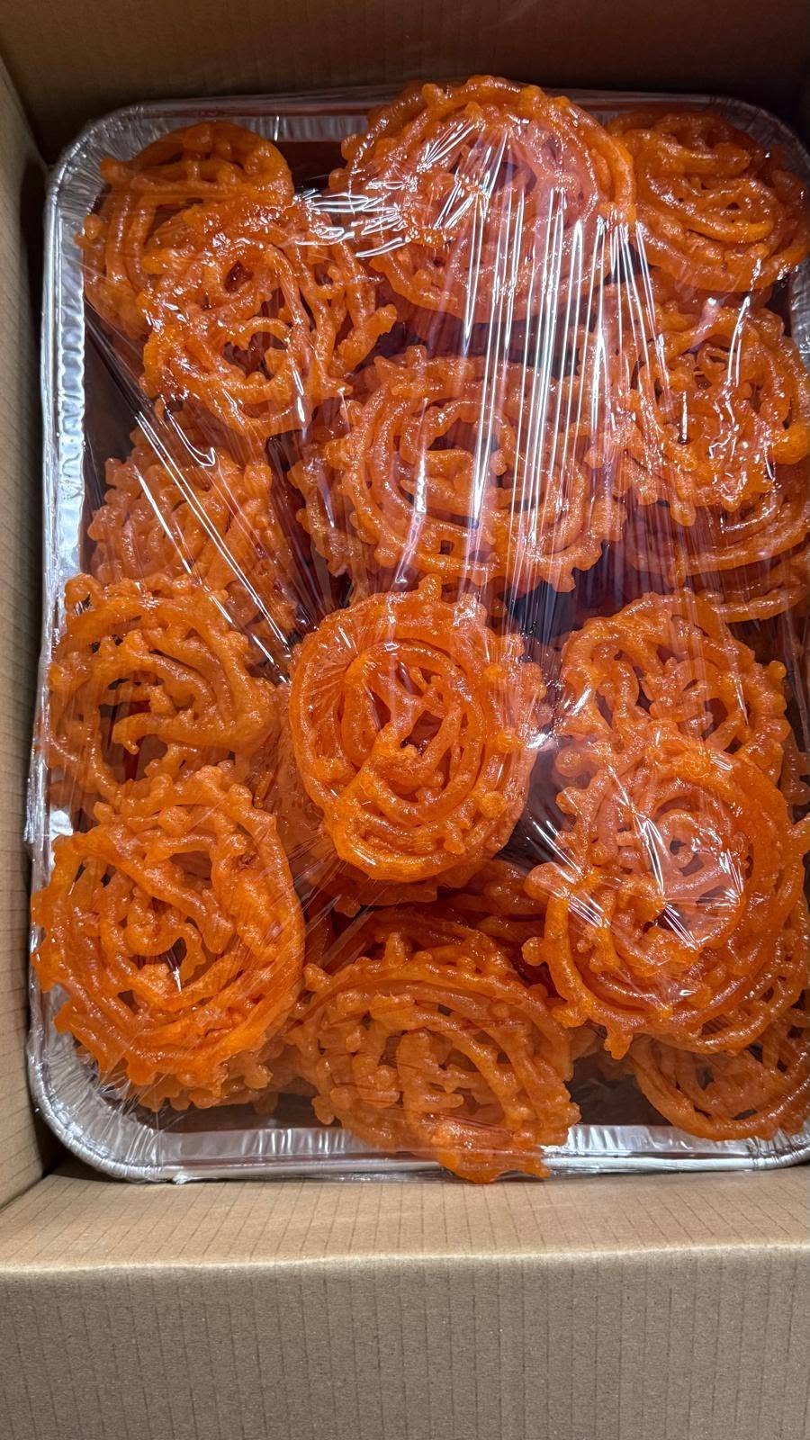 A foil tray of bright orange jalebi sweets under cling wrap
